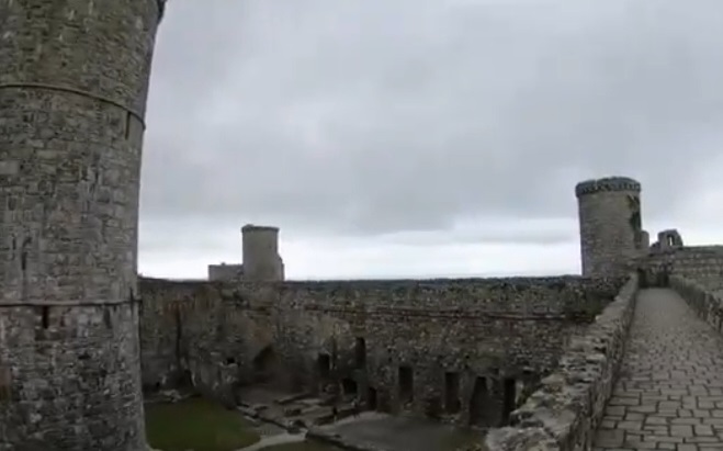 A view of Harlech Castle, highlighting its stone walls, towers, and the pathway along the battlements. The image shows the interior courtyard below, with remnants of medieval structures and a cloudy sky adding to the castle's dramatic and historic atmosphere